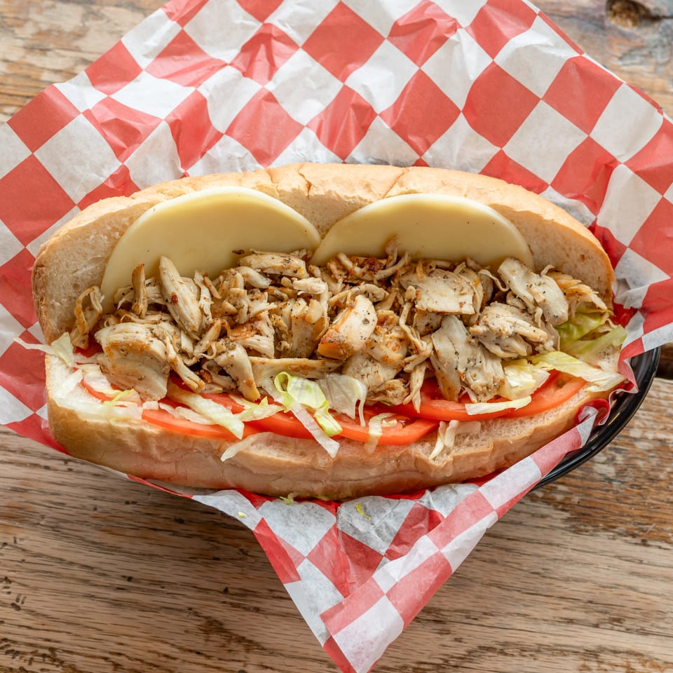 Chicken sub sandwich with cheese, lettuce, and tomatoes on red and white checkered paper.