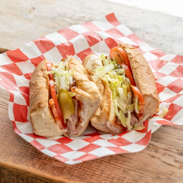 Two fully loaded submarine sandwiches with lettuce, tomatoes, and pickles on red and white checkered paper.