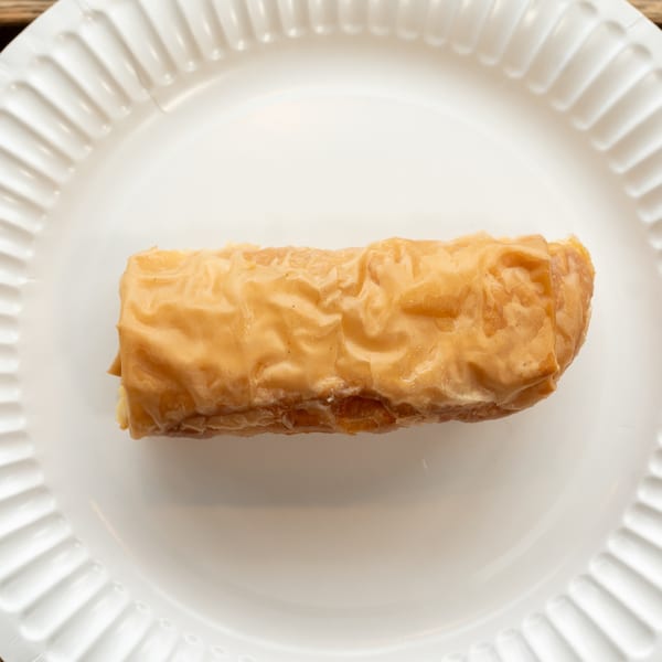 A golden-brown, rectangular baklava pastry, glistening with syrup, rests on a white paper plate.