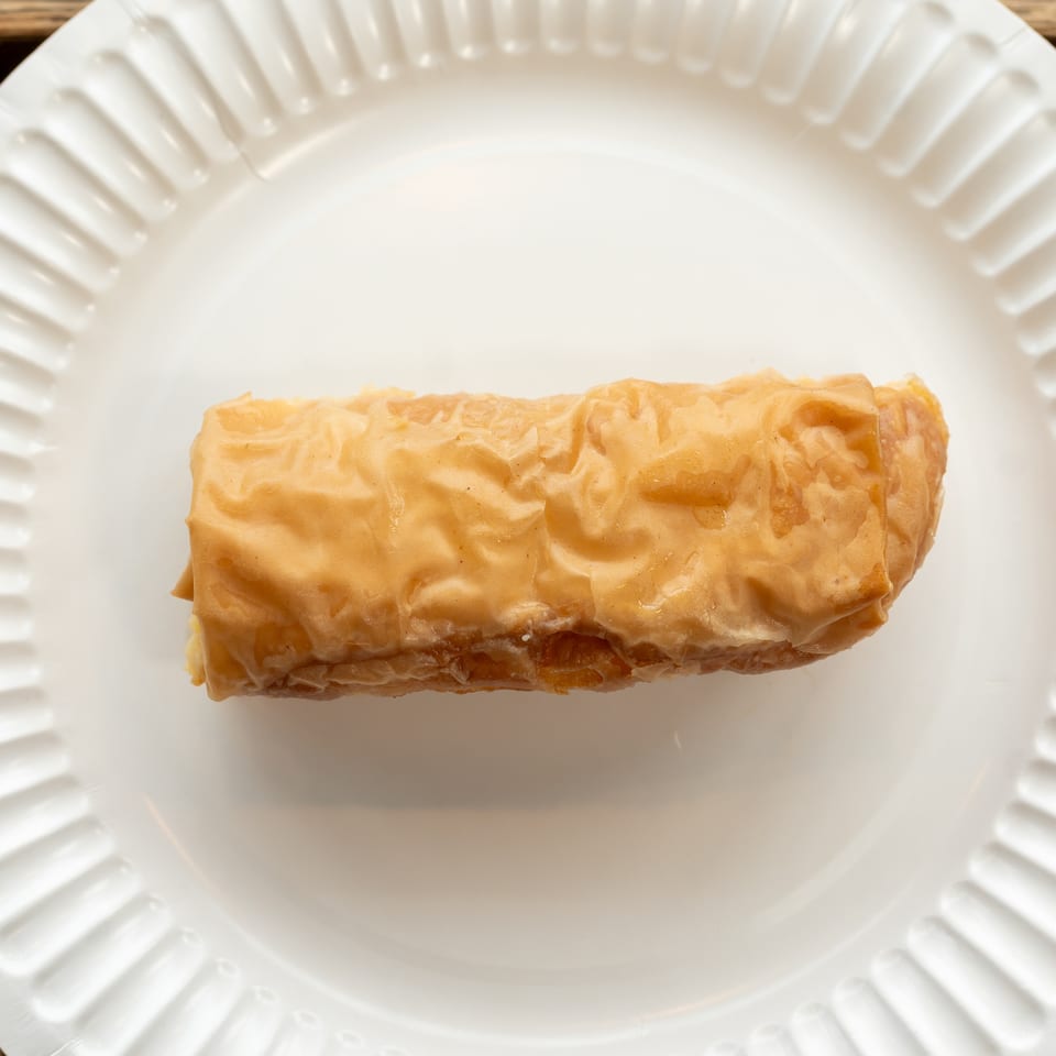 A golden-brown, rectangular baklava pastry, glistening with syrup, rests on a white paper plate.