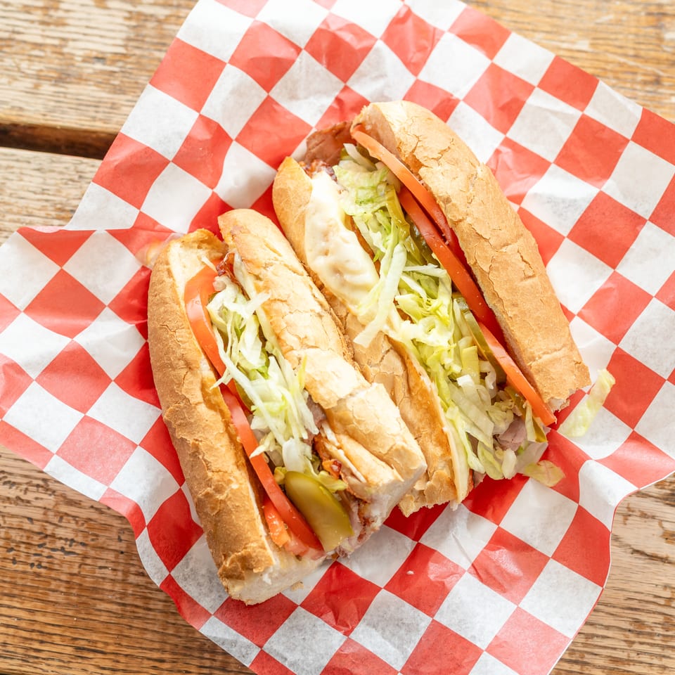 Two submarine sandwiches with meat, cheese, lettuce, tomatoes, and pickles on red and white checkered paper.